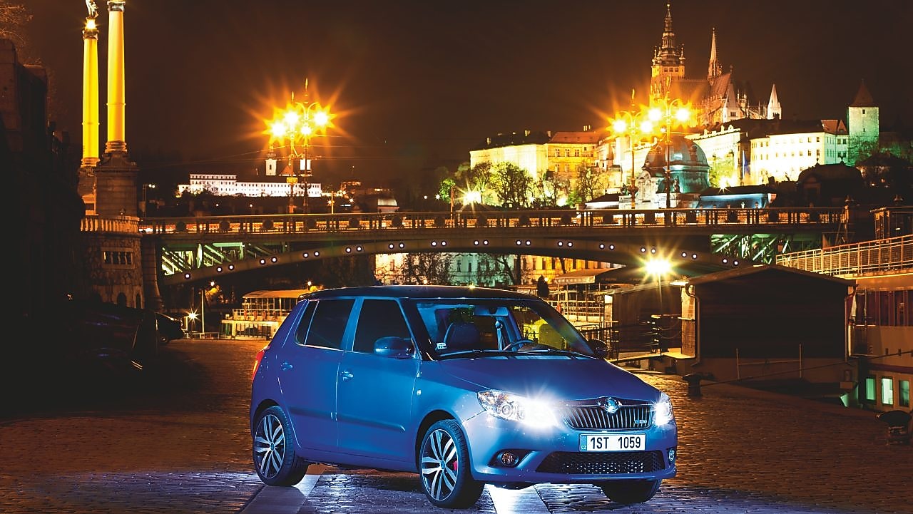 Blue car parked on a Prague road with a bridge and castle in the backdrop, at night