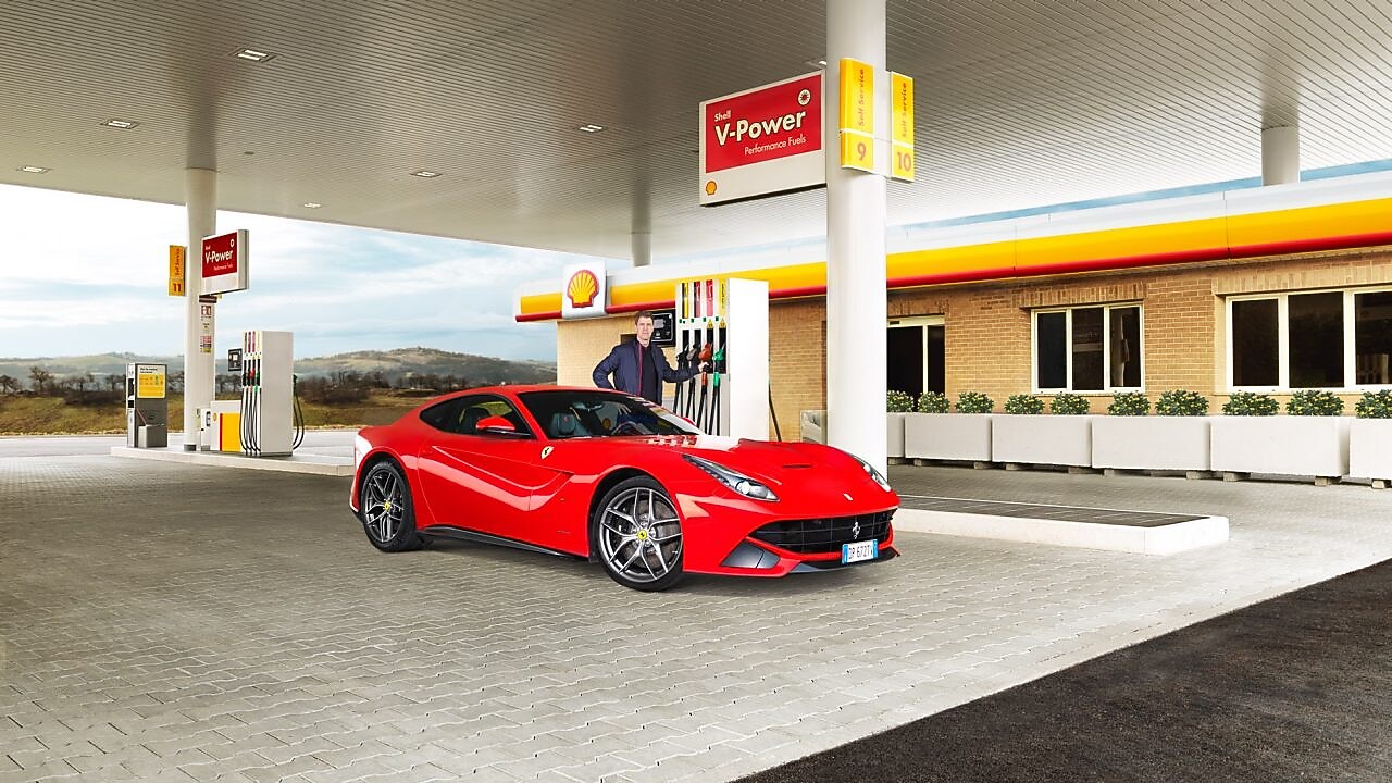 A red Ferrari sitting on a Shell station forecourt with a man leaning on a petrol pump